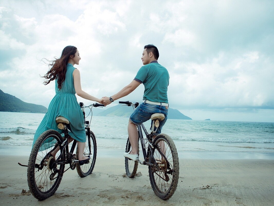 photo of young couple riding bikes and holding hands on the beach